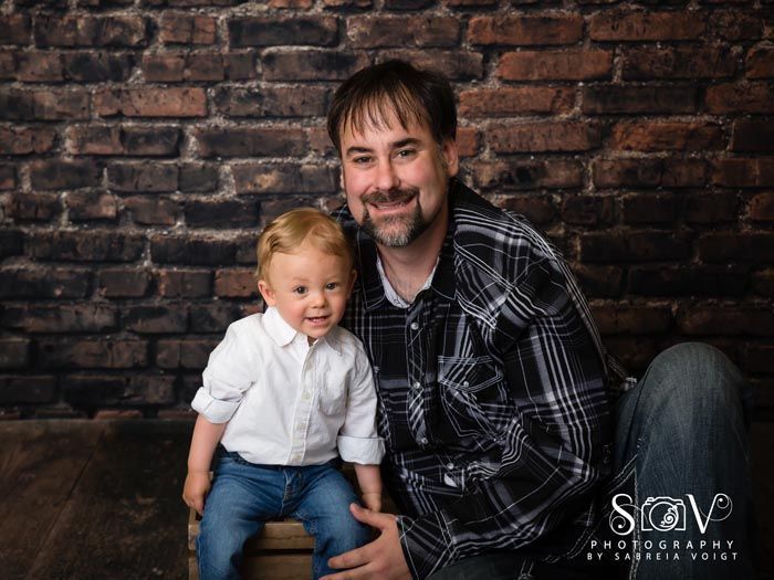 Man and young child smiling, posing in front of brick wall background.