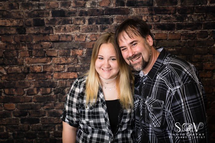 Woman and man smile, standing close, in front of a brick wall. Both wear plaid shirts.