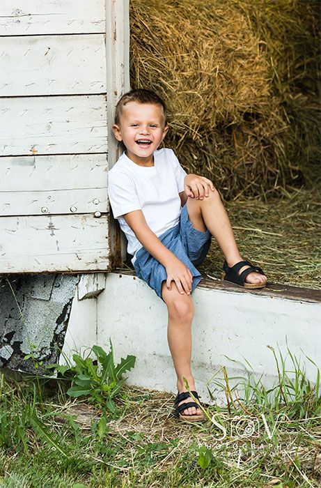 Boy in white shirt and blue shorts sits smiling at the opening of a barn with hay.