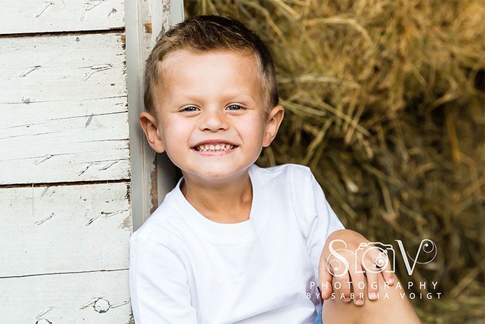 Smiling child in a white shirt, leaning against a white wooden wall, with hay in the background.