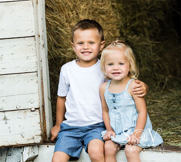 Boy in white t-shirt and girl in blue dress sitting close. Hay bales in the background.
