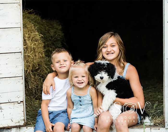Children with a black and white dog sit in a barn doorway, smiling.