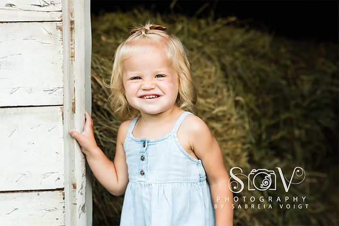 Smiling child in a blue dress stands in a doorway with a hay bale background.