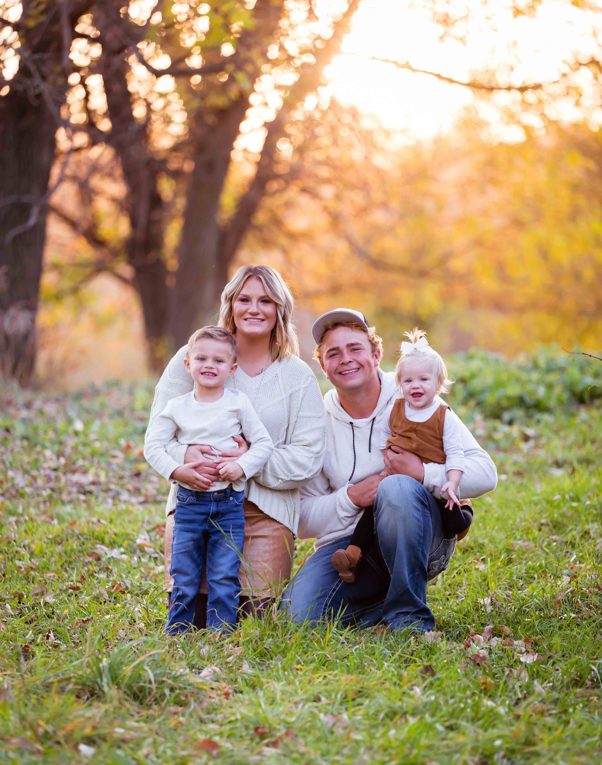 Family of four smiles for the camera in a grassy field with trees. Warm sunlight.