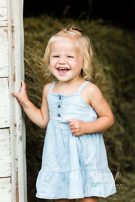 Smiling child in blue dress stands by a doorway, with hay in background.