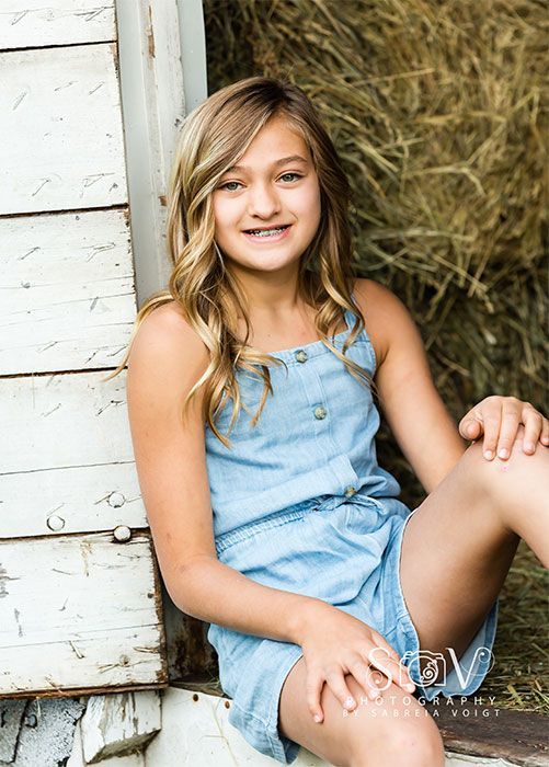 Girl in denim jumpsuit smiles, sitting near weathered wood and hay.