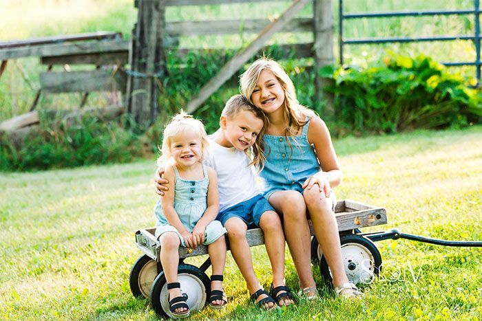 Three children smiling, sitting on a wagon in a grassy field.