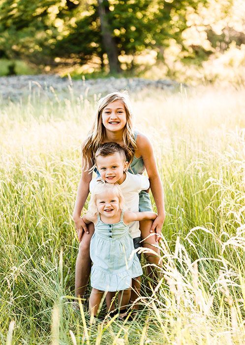 Three children in a grassy field smiling. Sunlight in background.