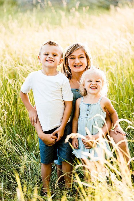 Three children smiling in tall grass. Boy in white shirt and shorts, girl in blue, blonde girl in blue dress.