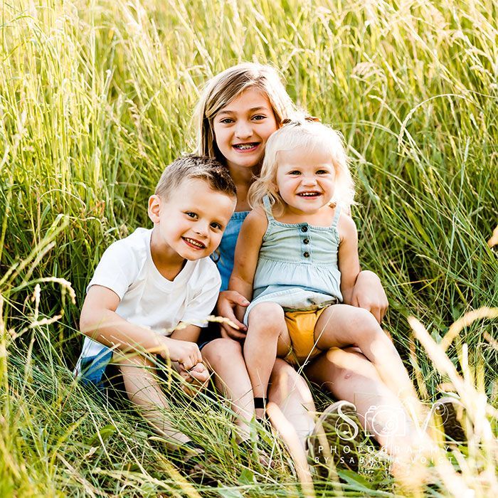 Three children smiling, sitting in tall grass. The older child is in the center with two younger children on either side.