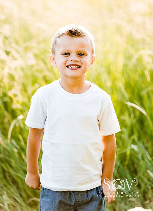 Smiling child in white shirt and blue shorts standing in tall grass, backlit by sunlight.