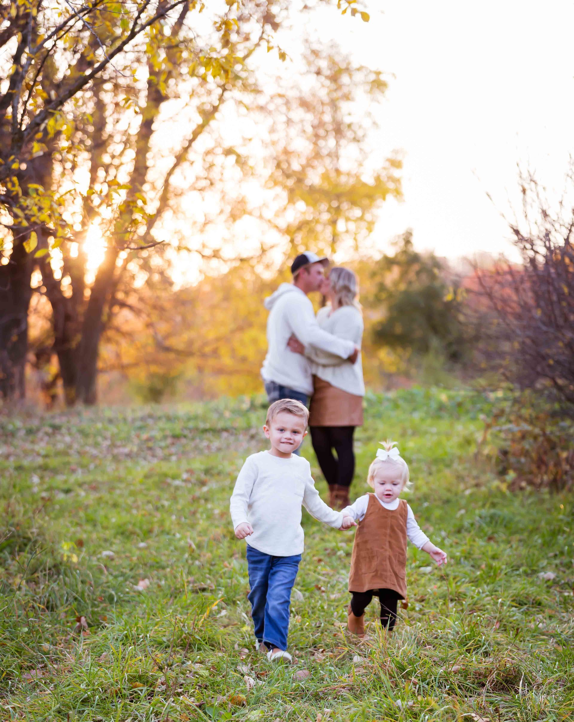 Family in park: parents hugging in background; two children walking in foreground, sunny autumn day.
