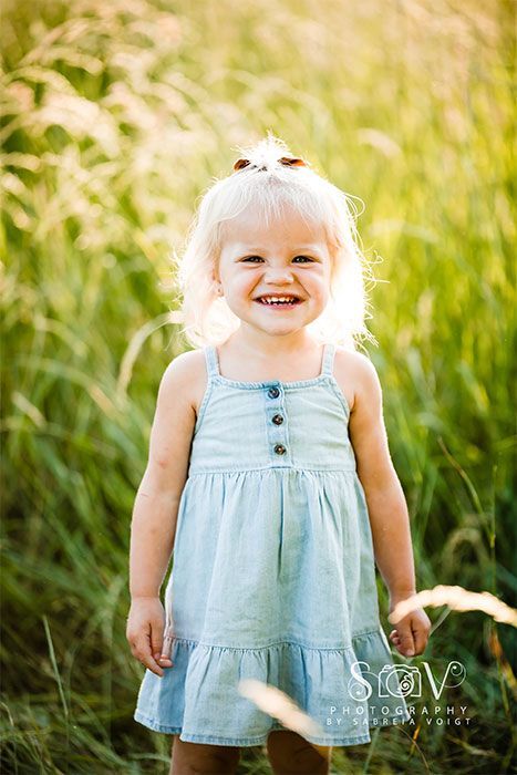 Blonde toddler in a blue sundress smiles in a sunlit field of tall grass.