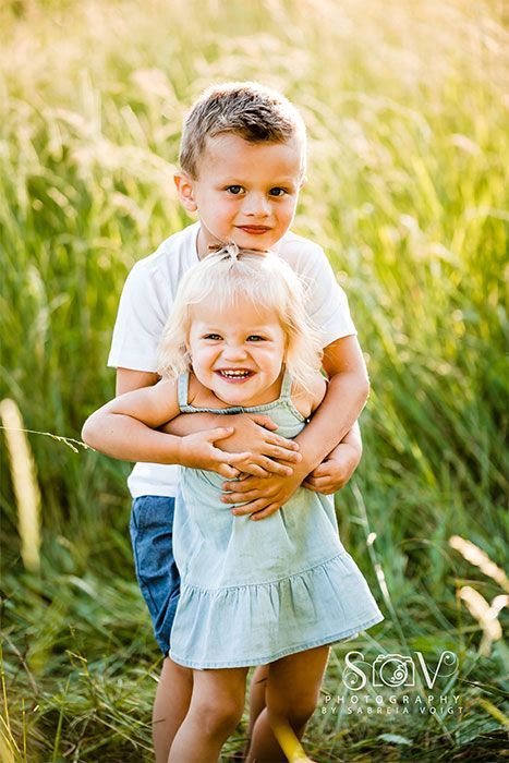 Boy hugging girl, both smiling in a field of tall grass. Sunlight streams, highlighting their faces and clothes.