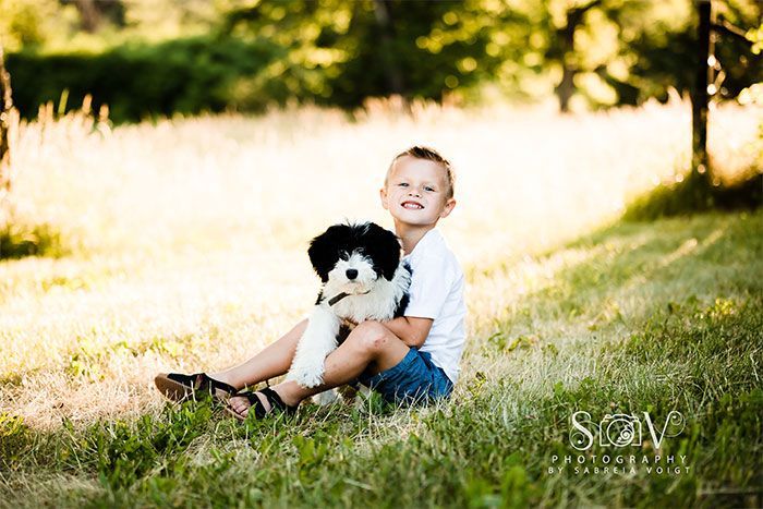 Boy in blue shorts holding a black and white puppy, sitting in grass, smiling.