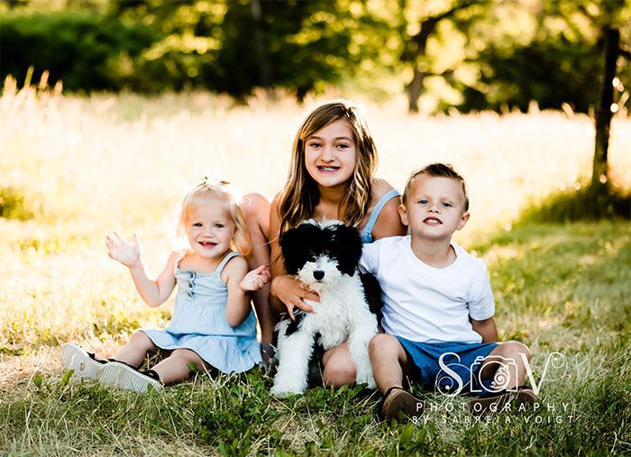 Three children and a black and white dog sit in a grassy field. Two children are smiling, one waving.