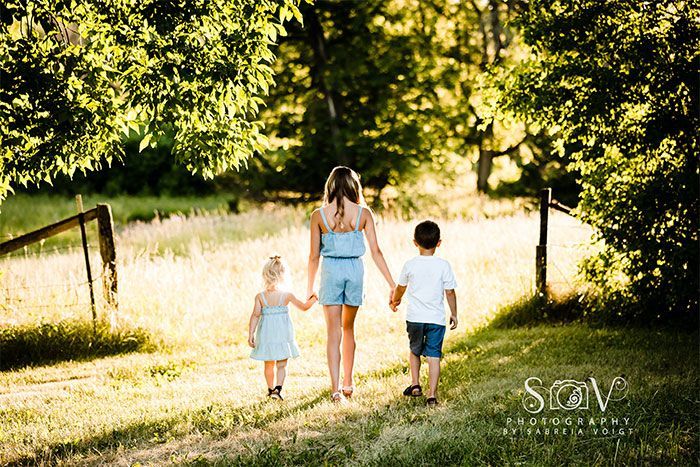Three children walk hand-in-hand toward a sunlit field, seen from behind.