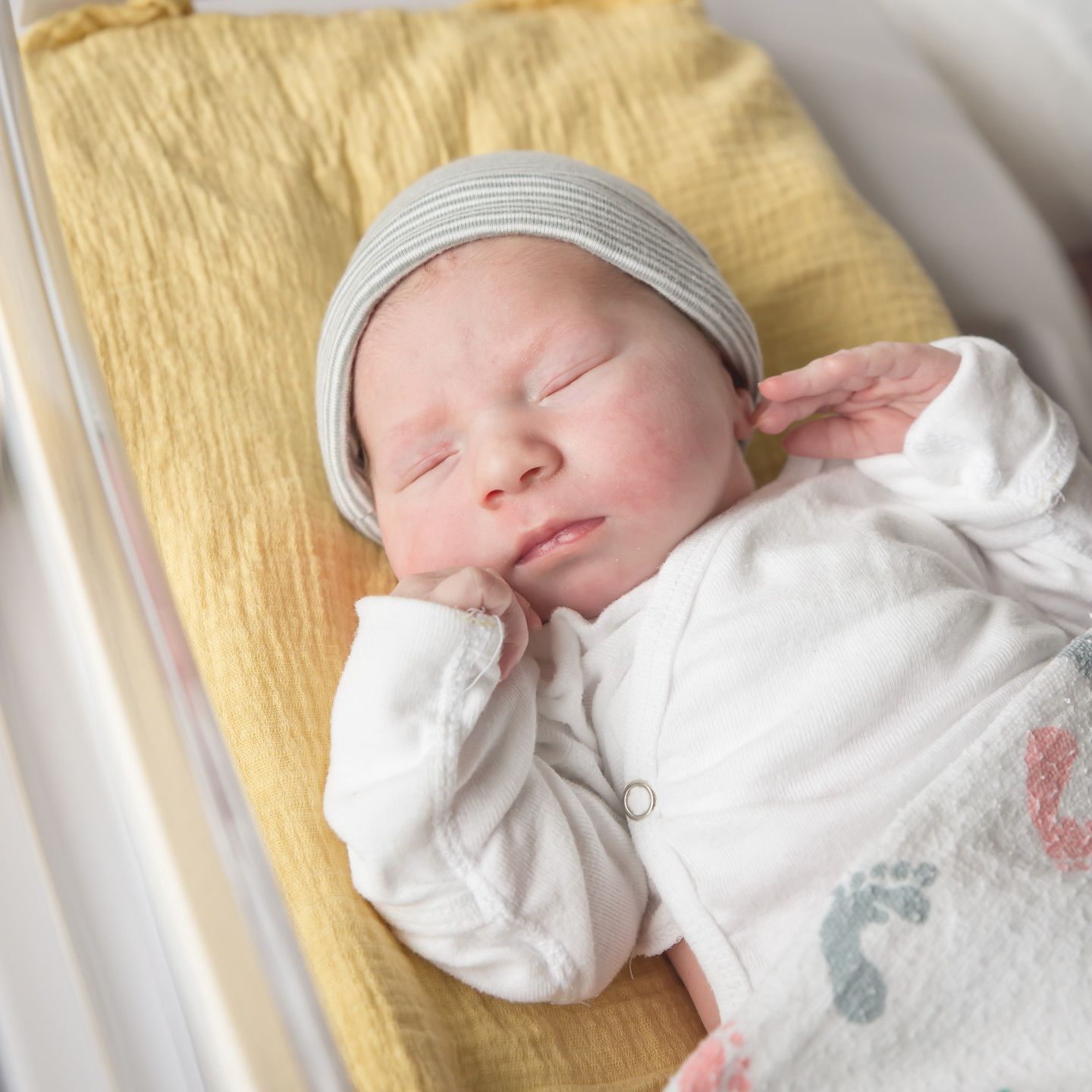 Newborn baby wearing a grey hat and white onesie, lying in a clear bassinet on a yellow blanket.