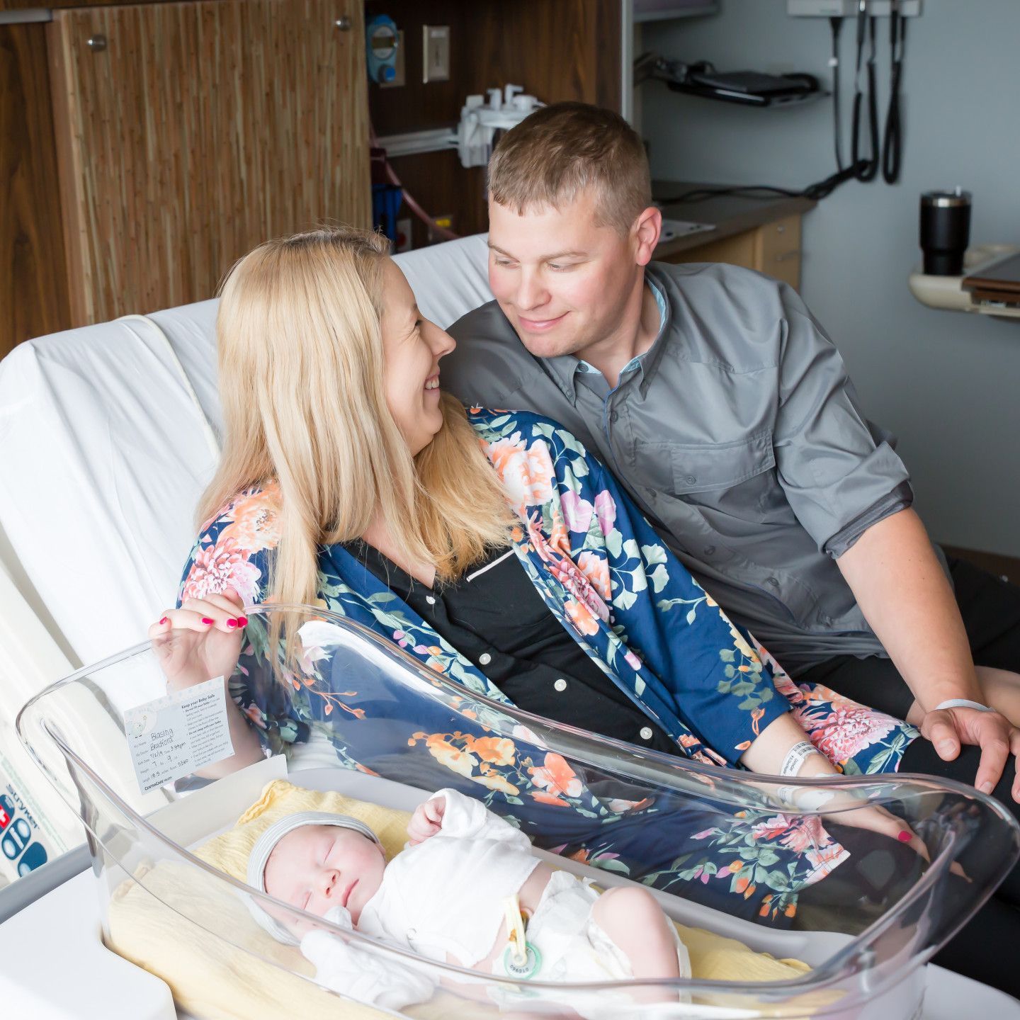 Parents gaze at newborn baby in hospital bassinet; woman in floral robe, man in gray shirt.