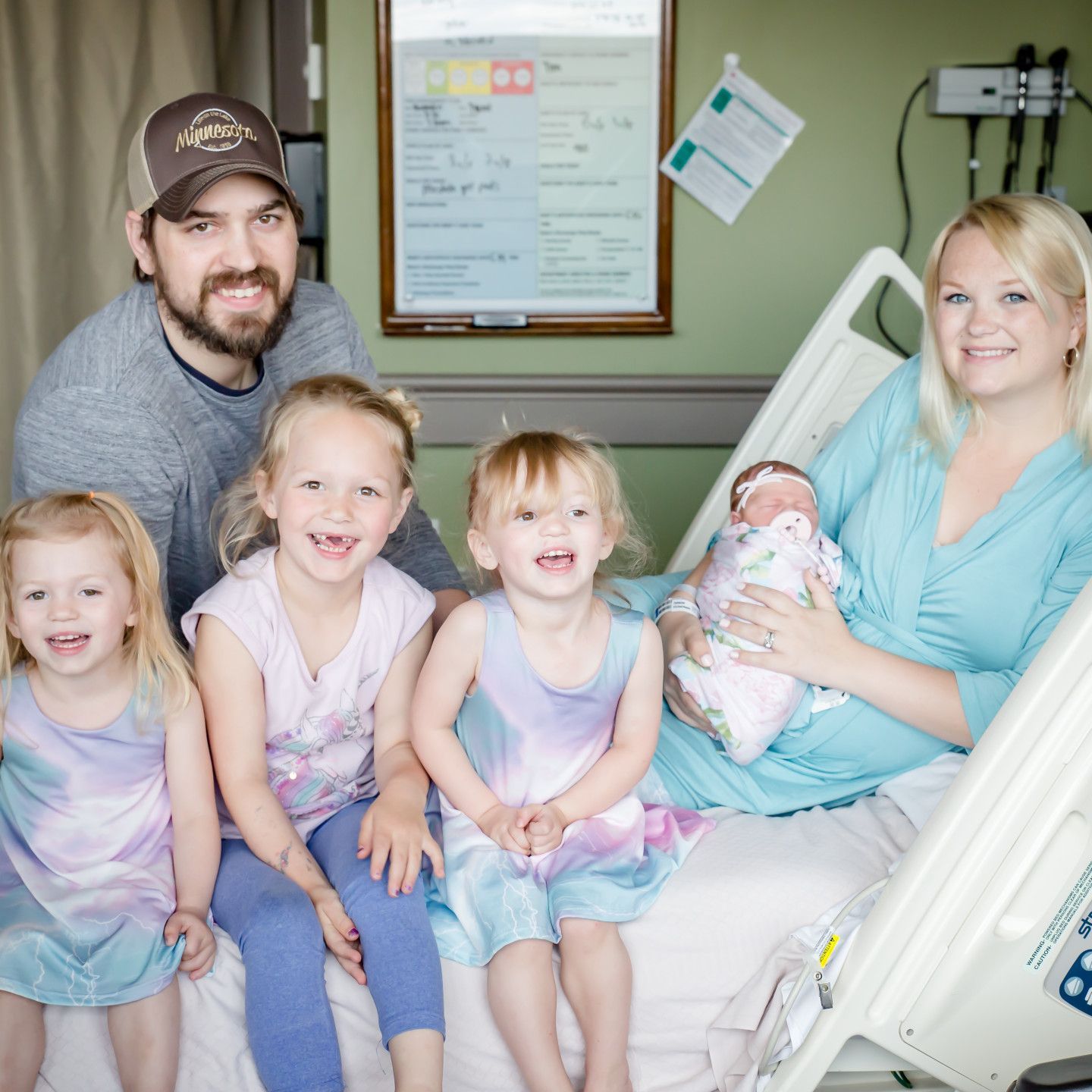 Family of seven poses in a hospital room; mother holds newborn, children smile, father looks at camera.