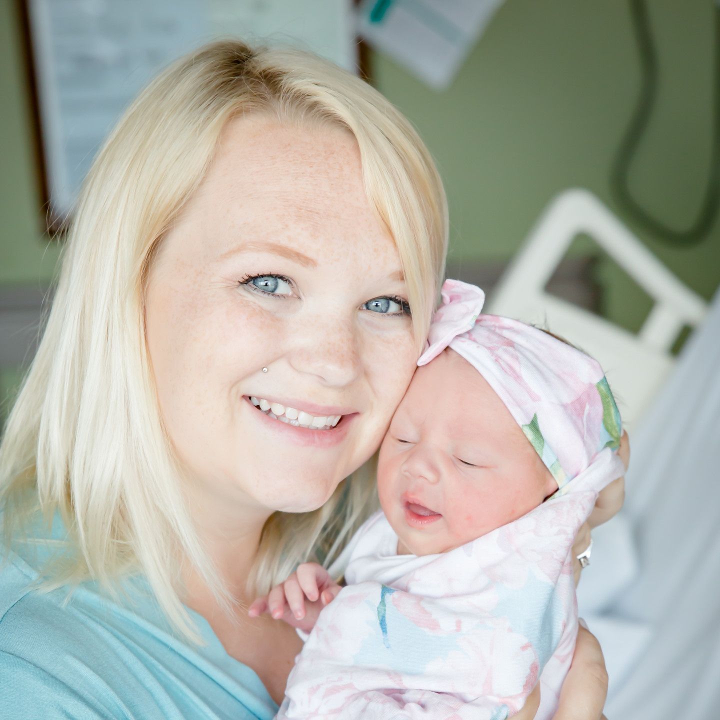 Woman with blonde hair smiles, holding newborn baby wrapped in a floral blanket in hospital.