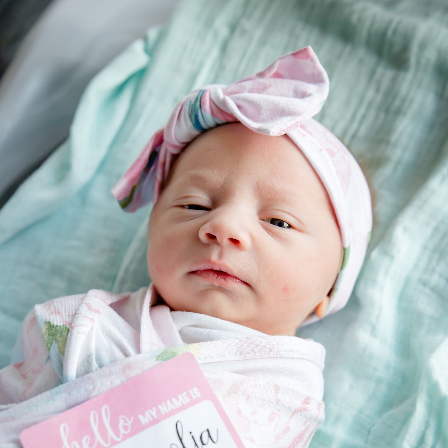 Newborn baby in floral wrap and pink bow headband, looking at the camera.