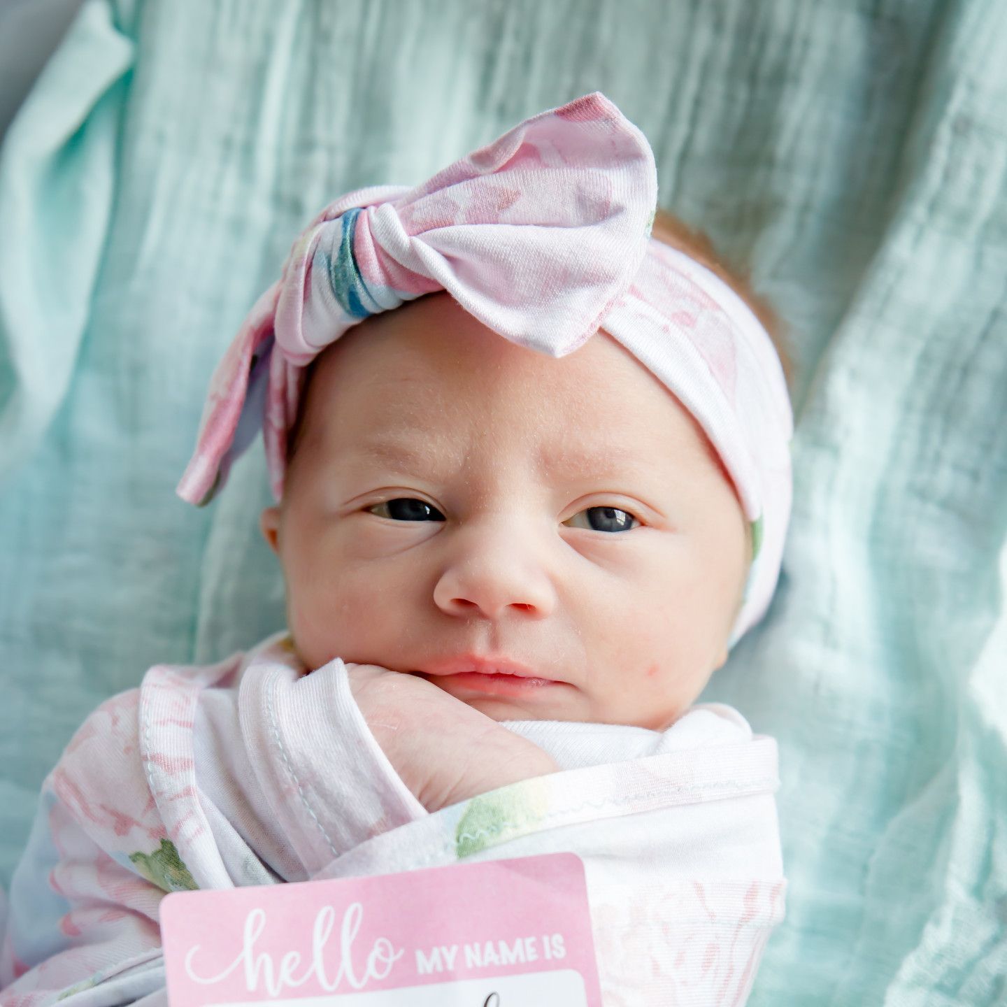 Newborn wrapped in a floral blanket, wearing a pink bow headband; holds a 