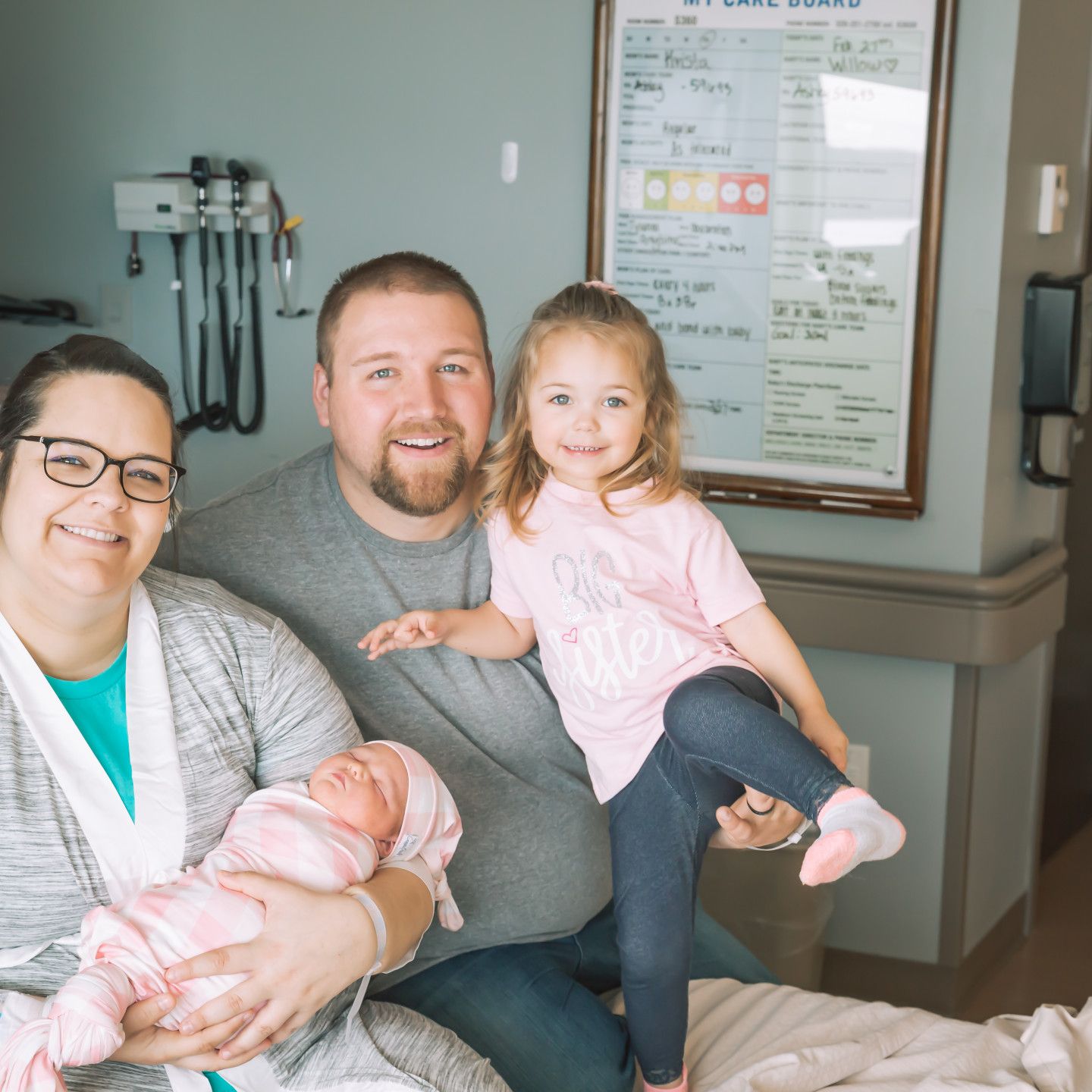 Family in hospital room with newborn. Parents smiling, older child posing, baby swaddled.