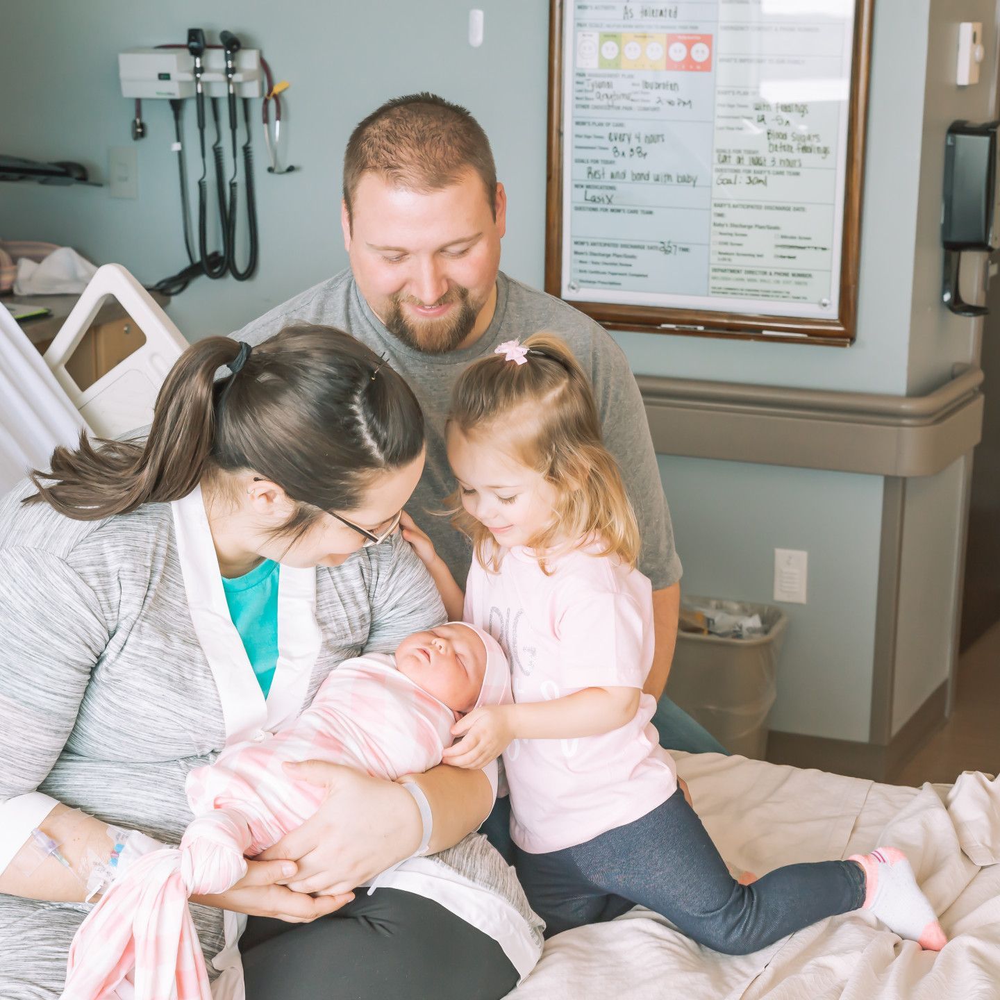 Family in a hospital room; mother holding newborn baby, father and older child smiling and looking at the infant.