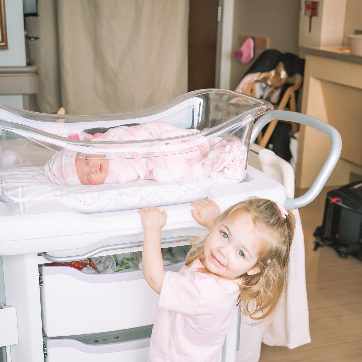 Young child smiles at camera while looking at newborn in hospital bassinet.