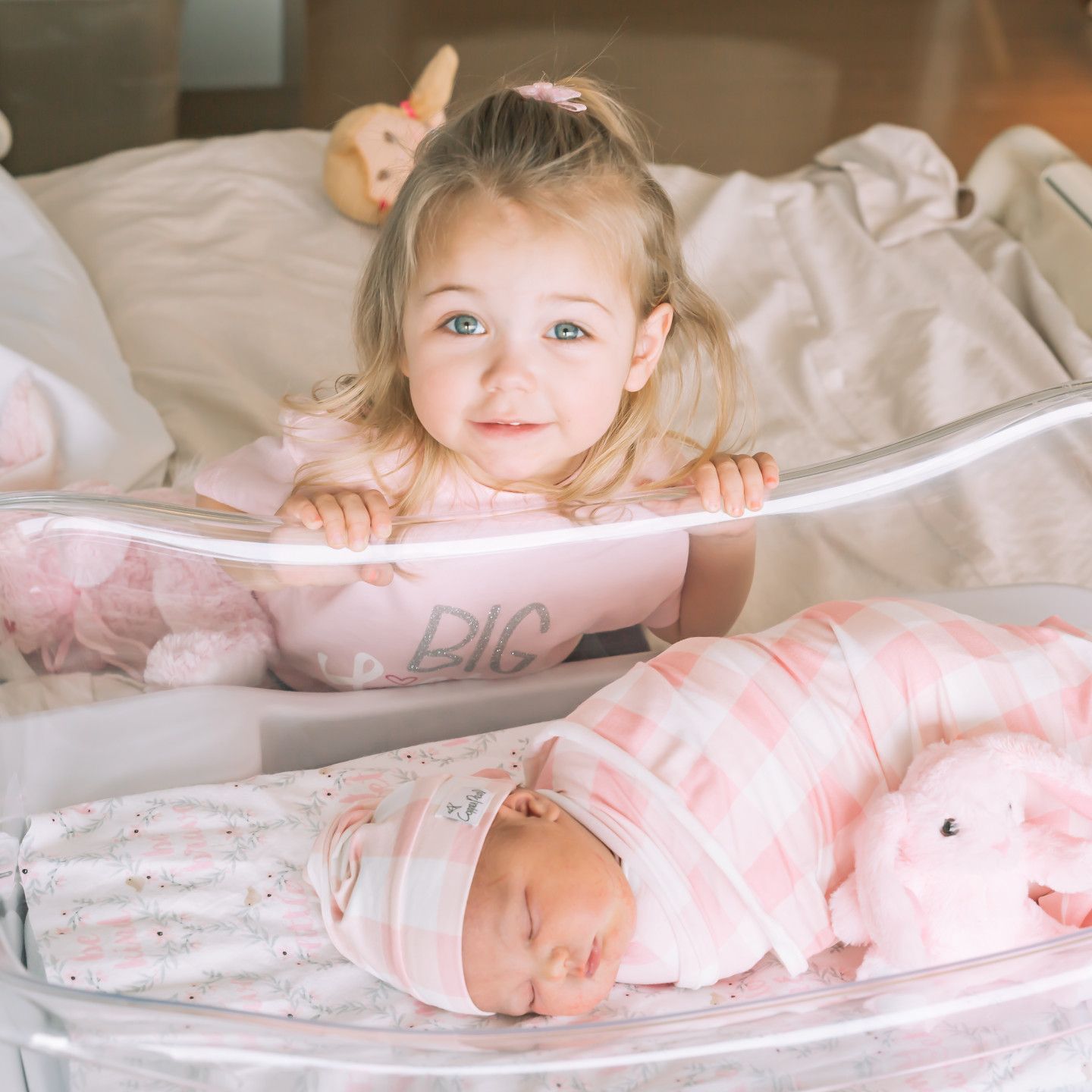A toddler smiles at the camera, looking over a bassinet with a sleeping newborn baby inside, all in pink.