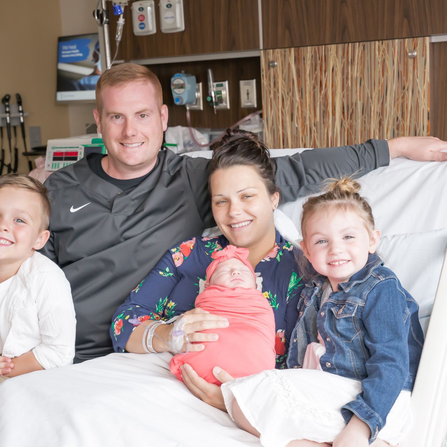 Family smiles while gathered in a hospital room, holding a newborn baby wrapped in a pink blanket.