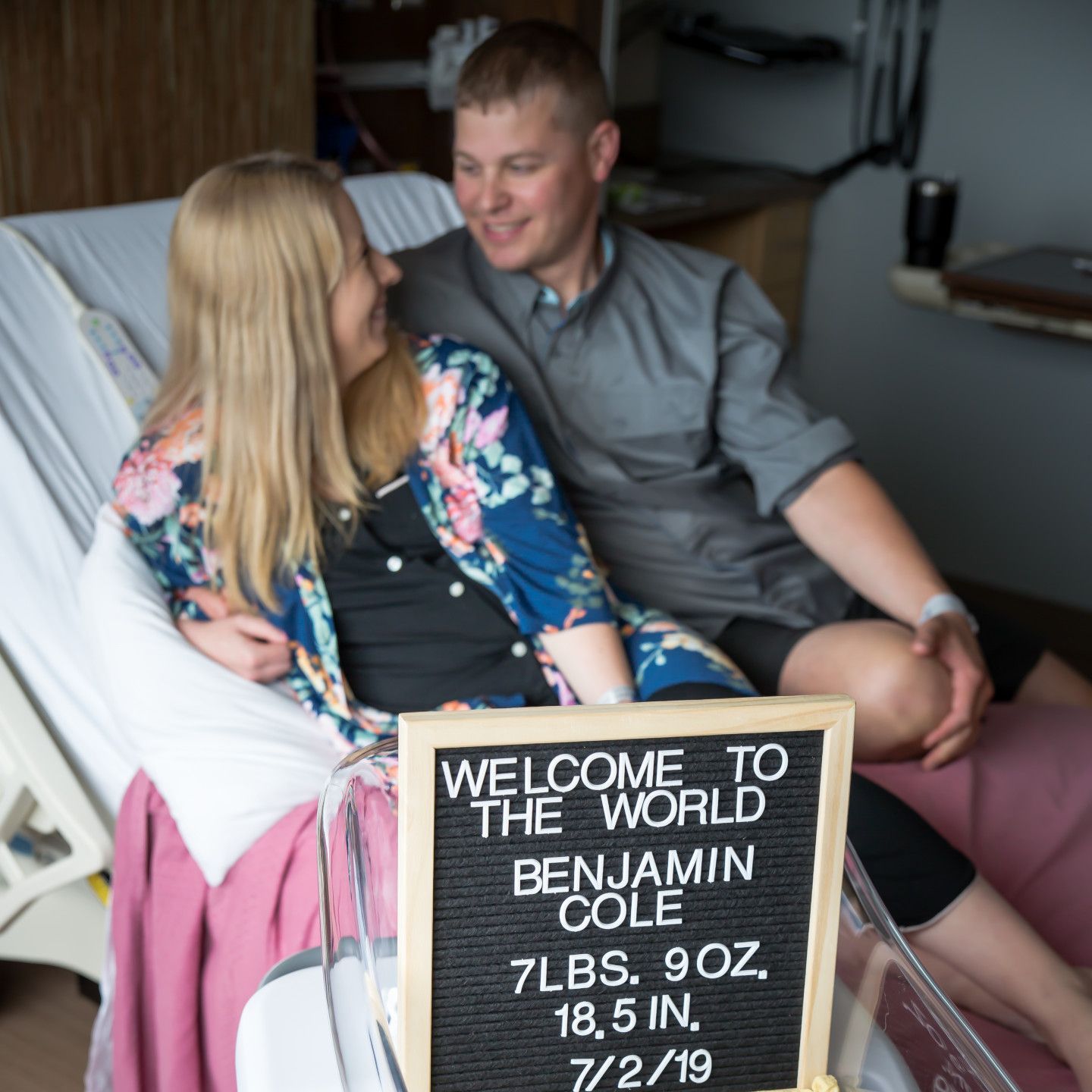 New parents in a hospital room smile at each other, holding their newborn's announcement sign.