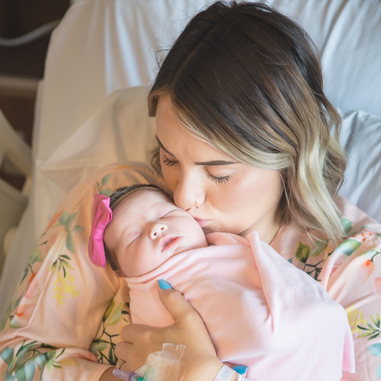 Woman in hospital bed kisses newborn baby wrapped in pink blanket.