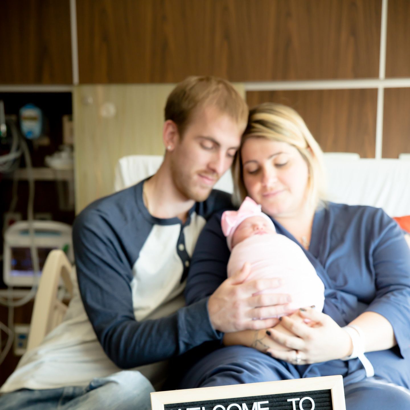 Parents holding newborn baby in hospital room; mom in robe, dad in long-sleeved shirt; baby wrapped in pink.