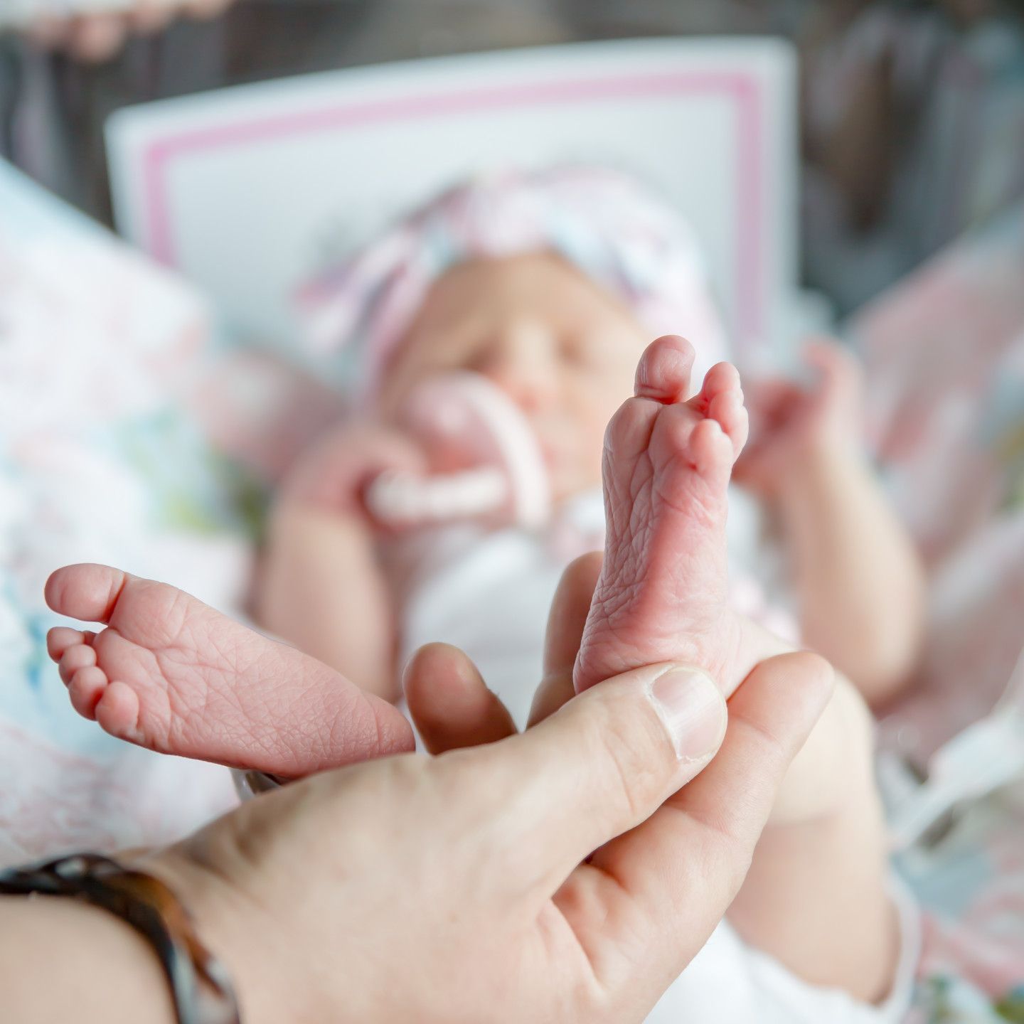 Newborn baby's tiny feet held in a person's hand. Baby is wearing a pink headband and lying in a crib.