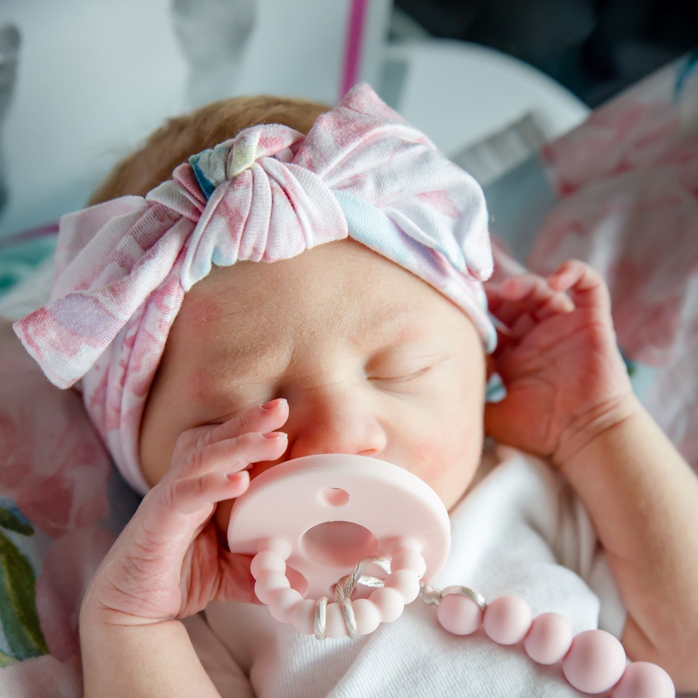 Newborn baby wearing a pink headband and using a pacifier, lying down.
