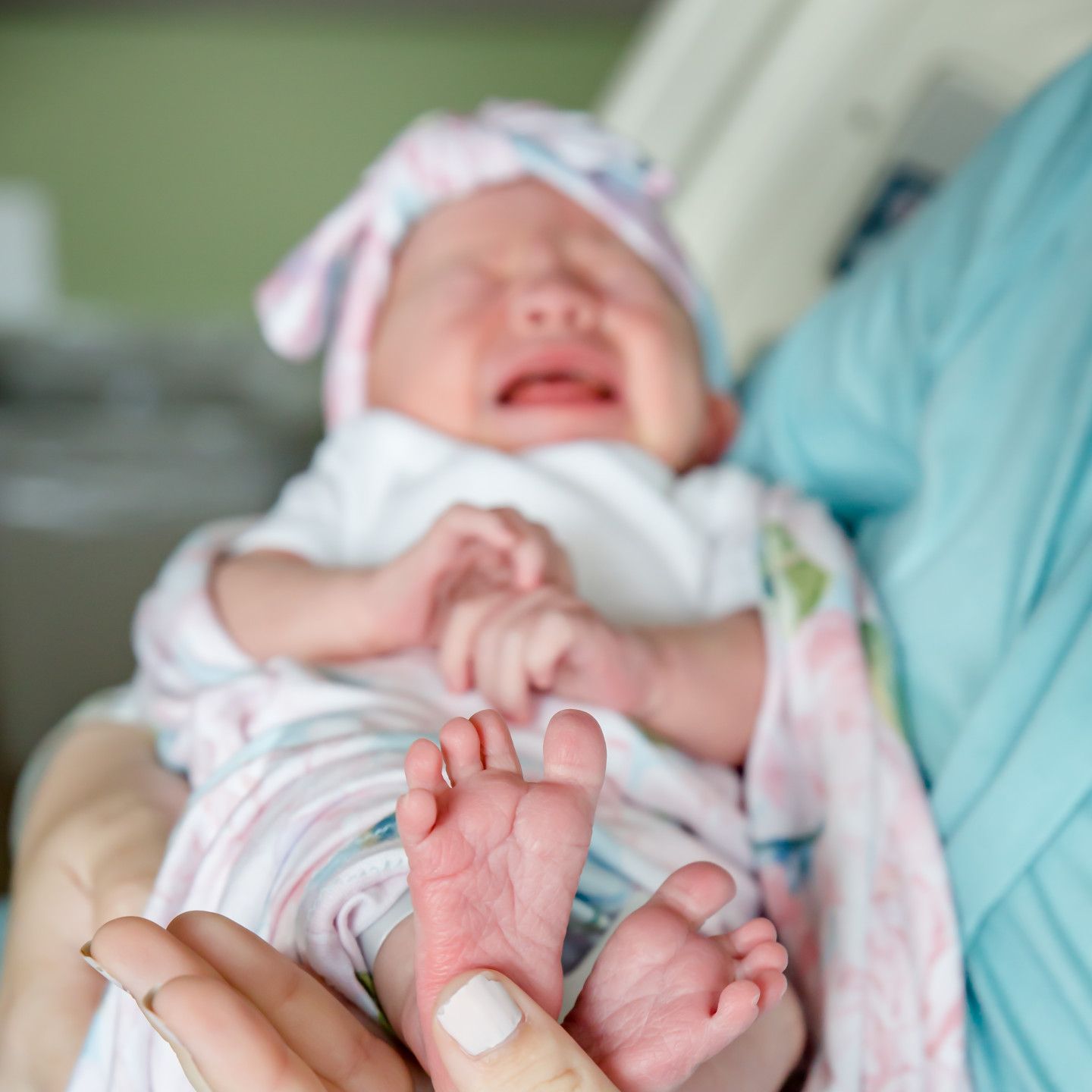 Newborn baby wrapped in blanket, held by a person. Baby is crying with closed eyes.
