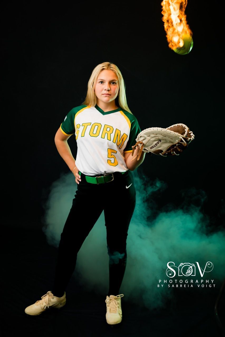 Softball player in uniform, holding glove, ball on fire, green and black background.