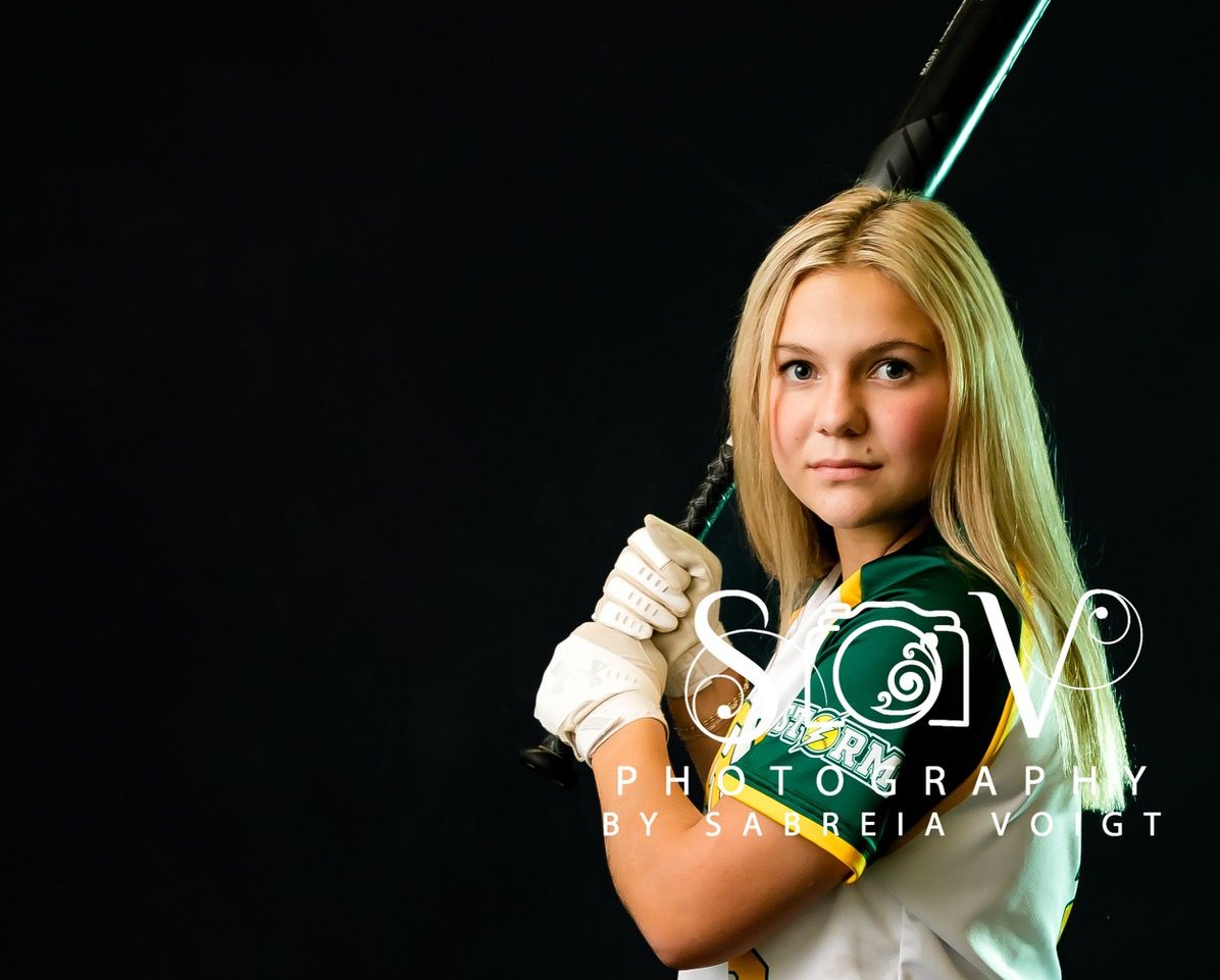 Blonde softball player holding a bat against a dark background, wearing a green and white uniform, looking forward.