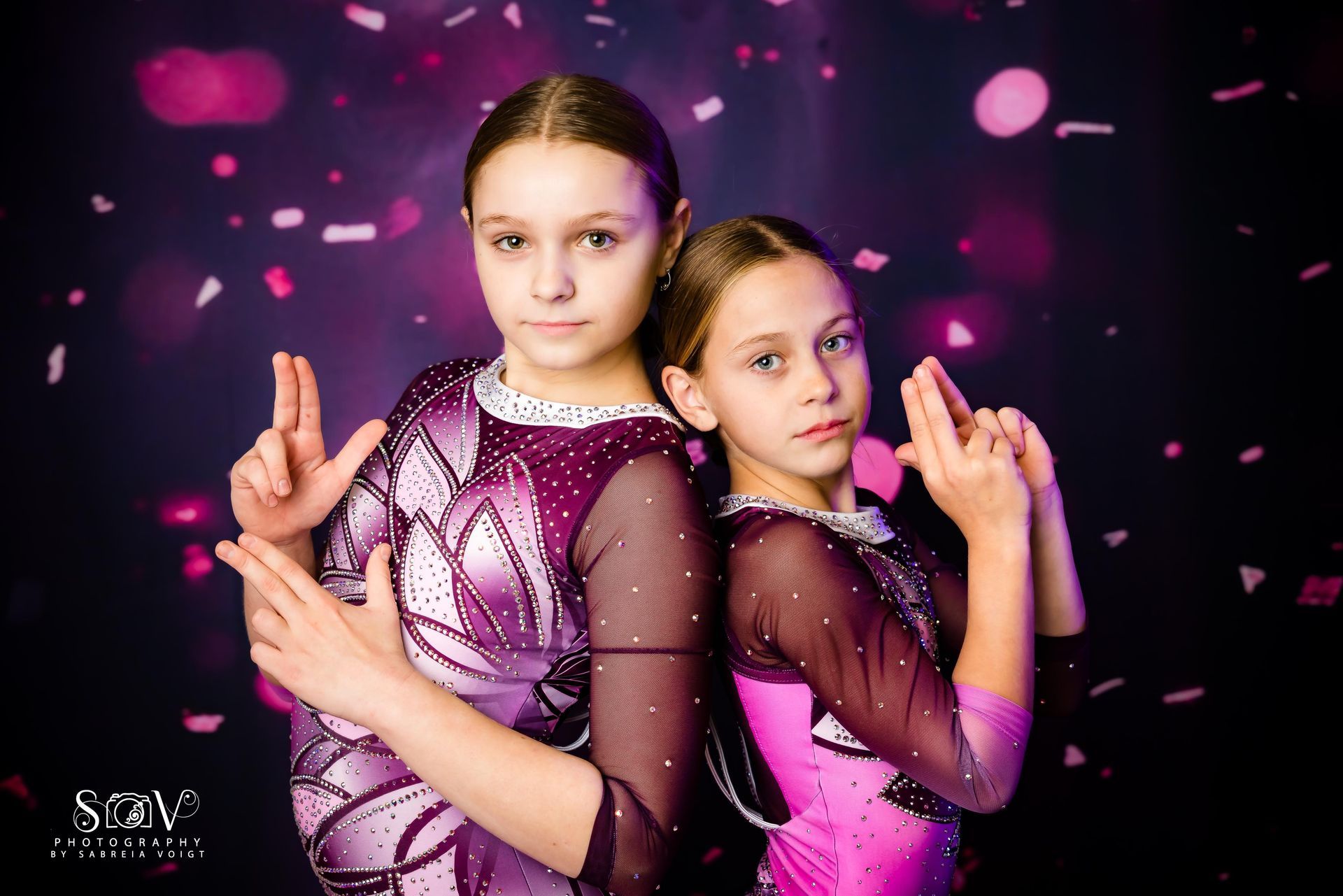 Two girls in matching purple leotards posing, making hand gestures, against a pink confetti background.