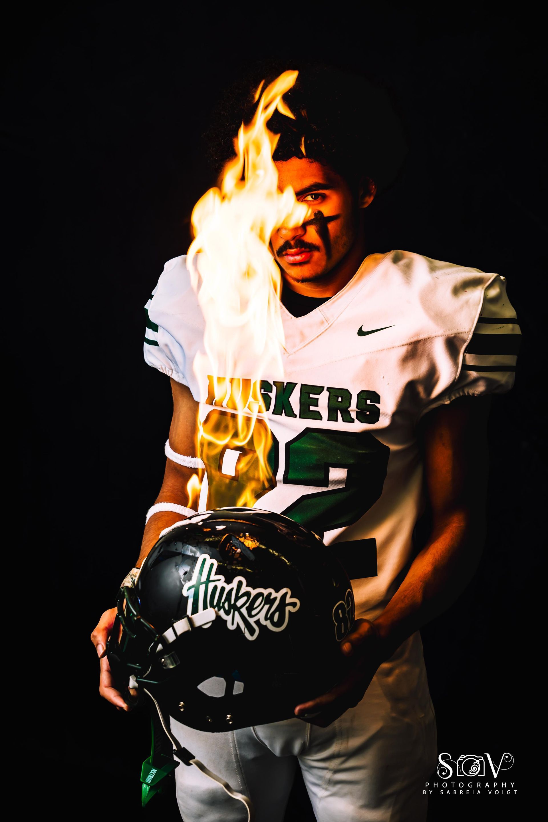 Football player in uniform, flame on head, holding helmet. Black background.