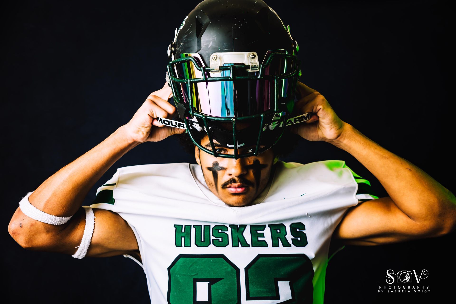 Football player in white and green Huskers jersey adjusting black helmet with green visor.