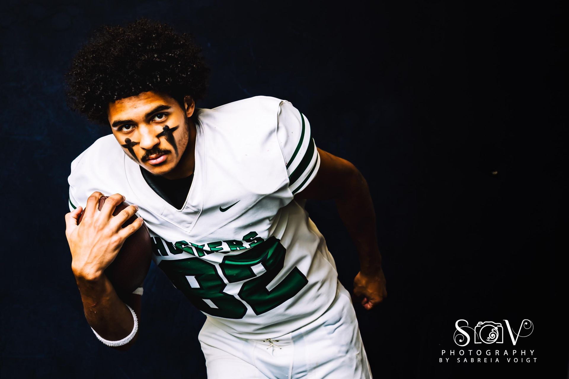 Football player in white and green jersey with face paint, holding a football.