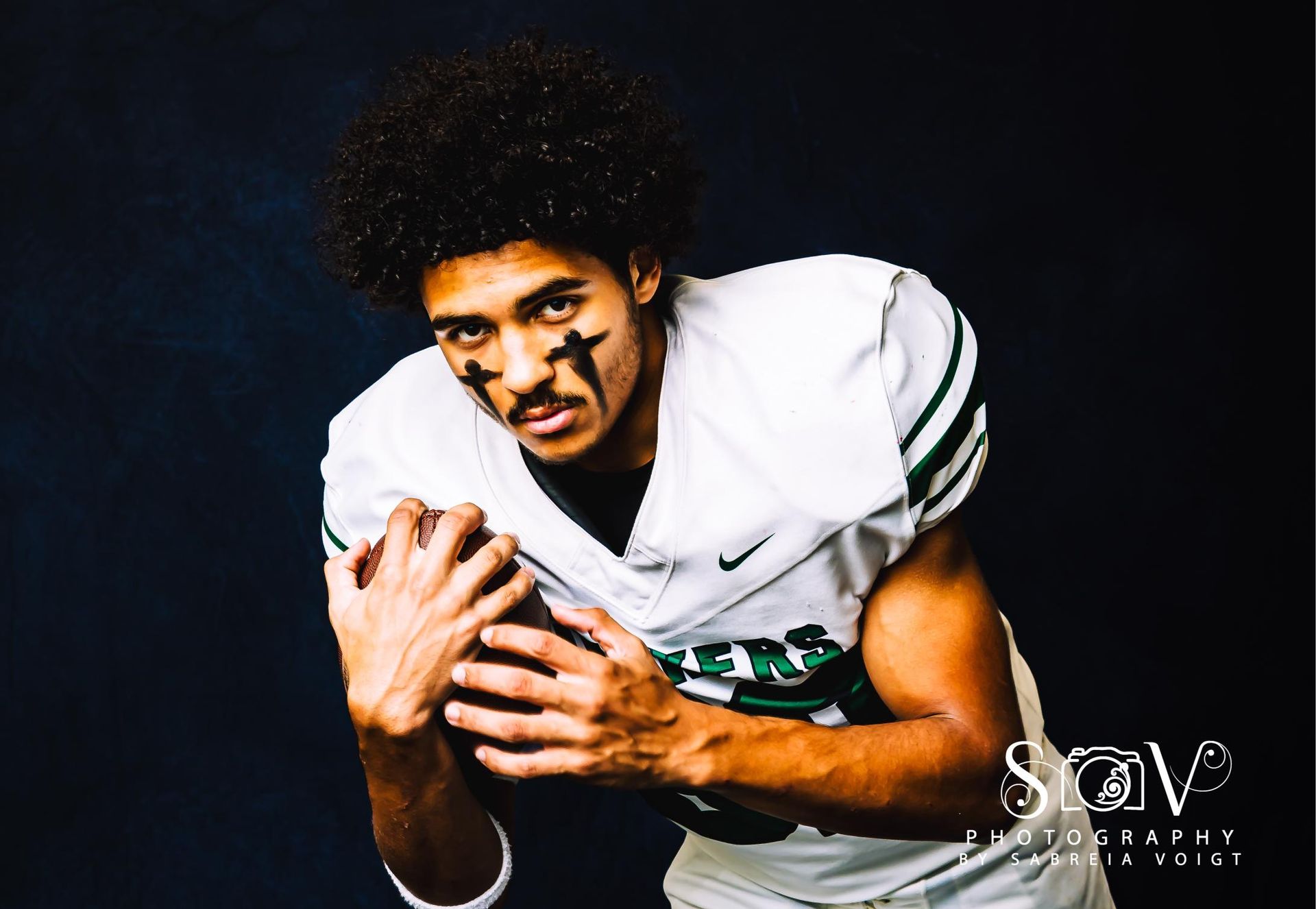 Football player in a white and green jersey with eye paint, holding a football against a dark blue background.