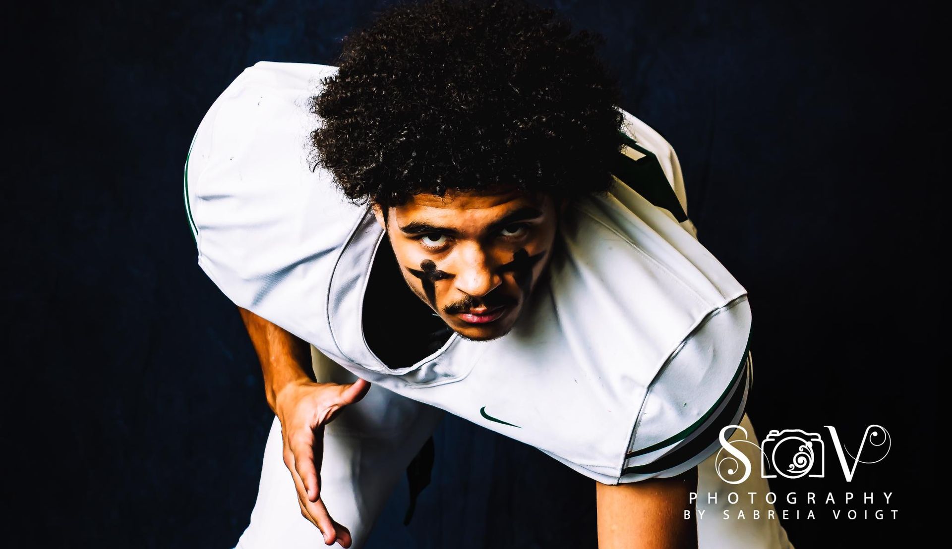 Football player in white and green uniform, face paint, in a crouched stance, looking forward.