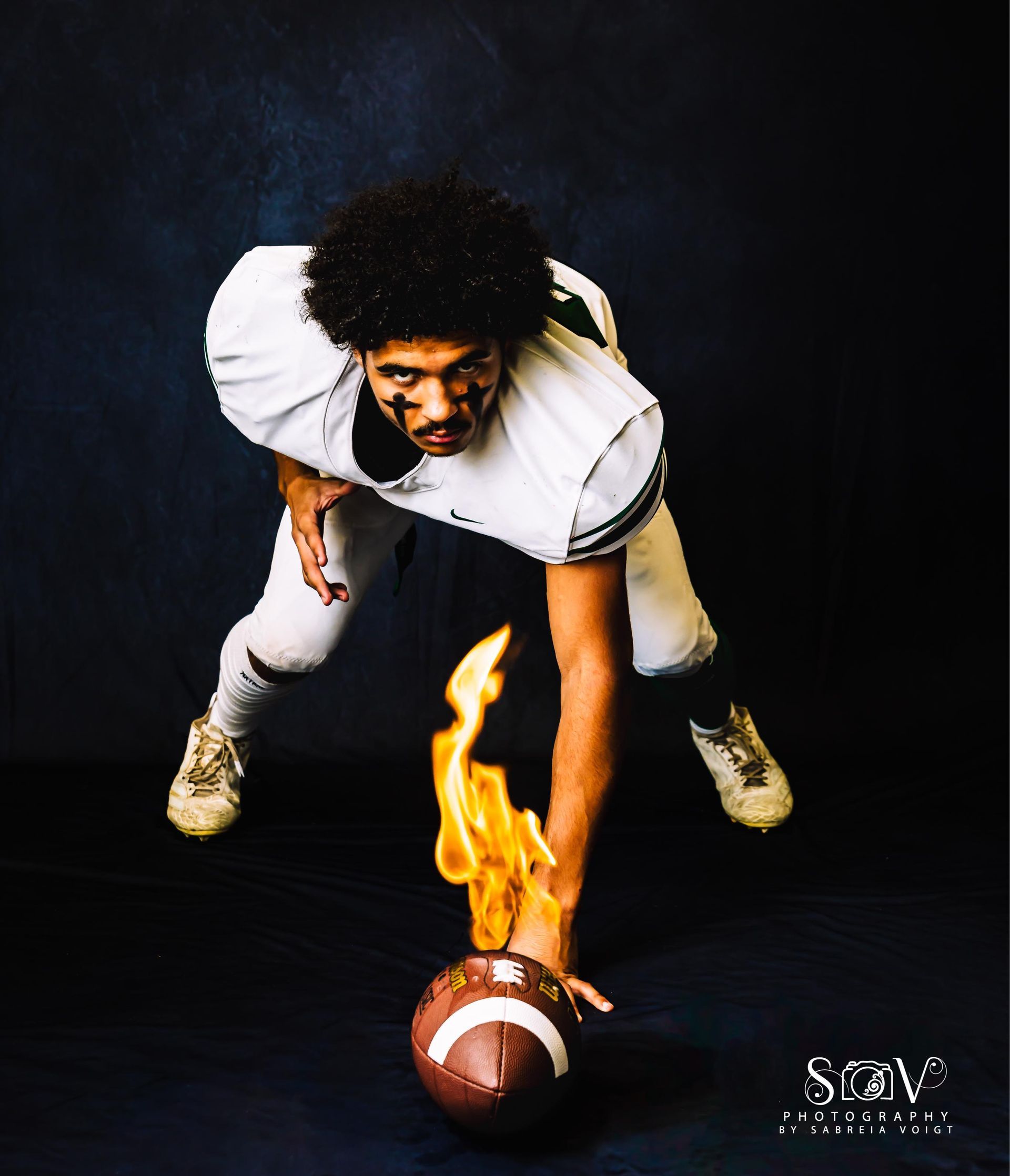 Football player in uniform reaching for a flaming football against a dark blue background.