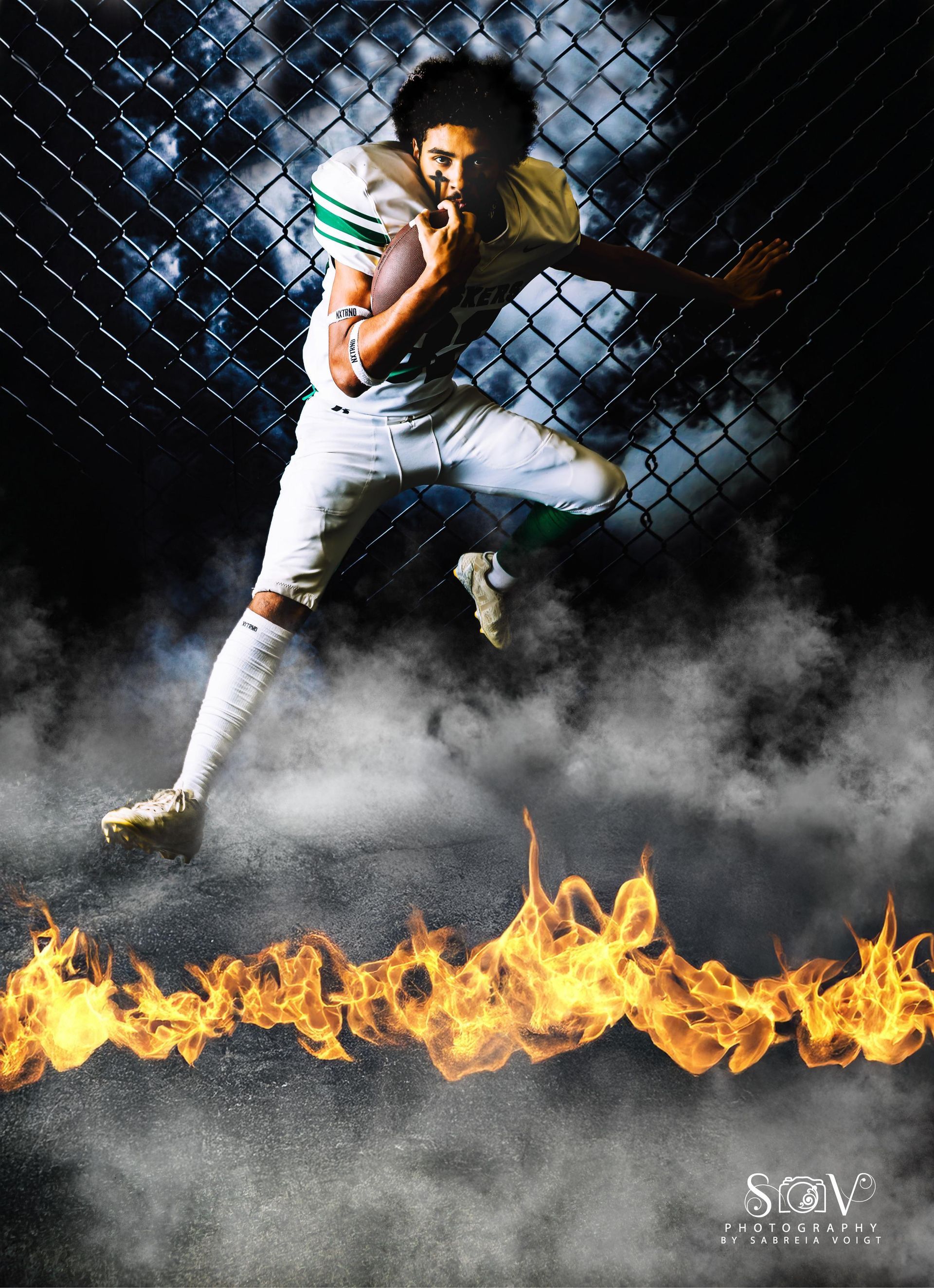 Football player in white uniform leaping, holding ball. Flame and smoke effect against chain link fence.