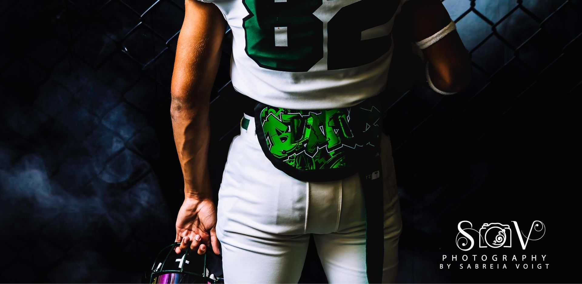 Football player in white uniform with green accents, holding helmet. Dark background with smoke.