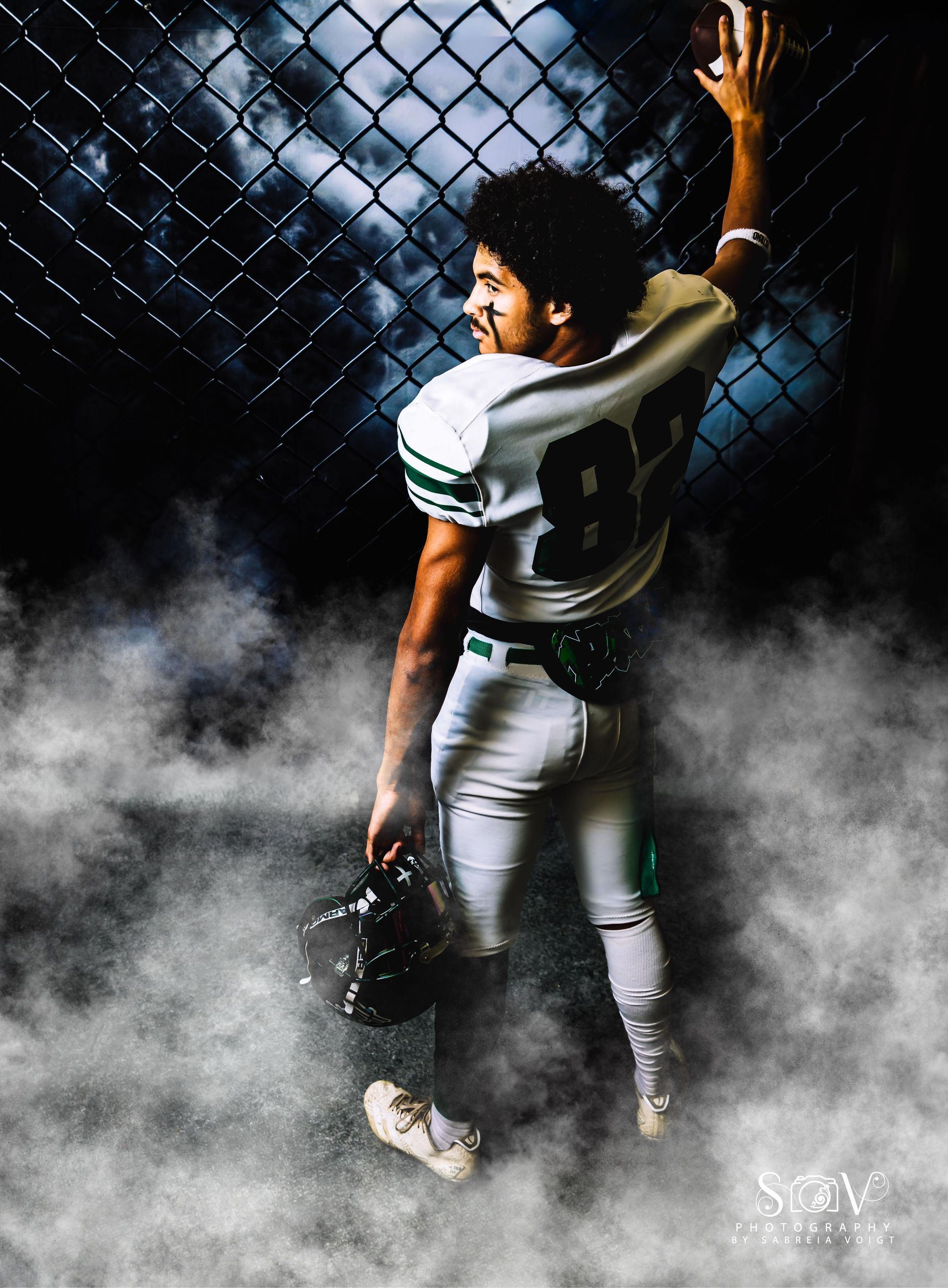 Football player in white and green uniform, holding helmet, looking away, next to a chain link fence, surrounded by fog.